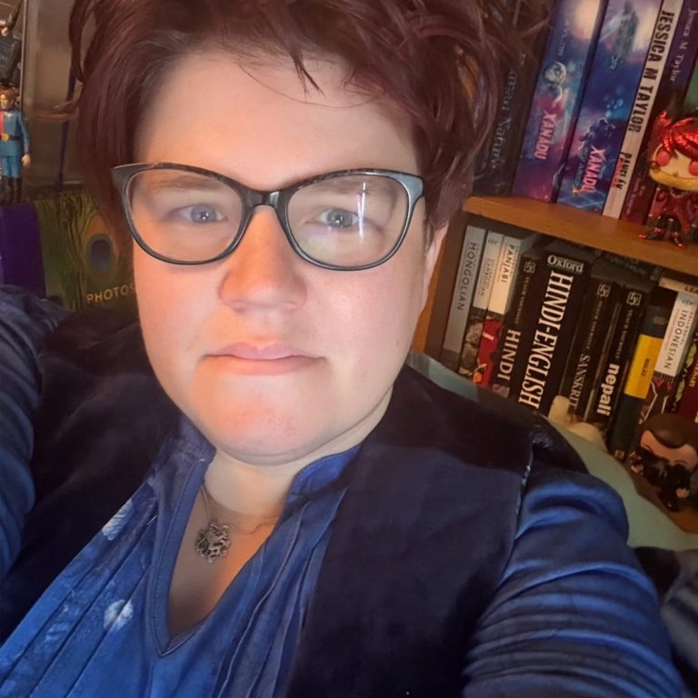 Photograph of a dark haired woman with glasses sitting in front of a bookcase with an Audrey II Funko in the background