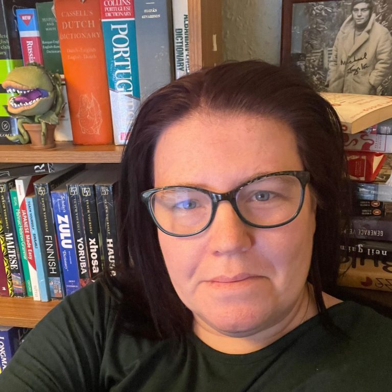 Photograph of a dark haired woman with glasses sitting in front of a bookcase with an Audrey II Funko in the background